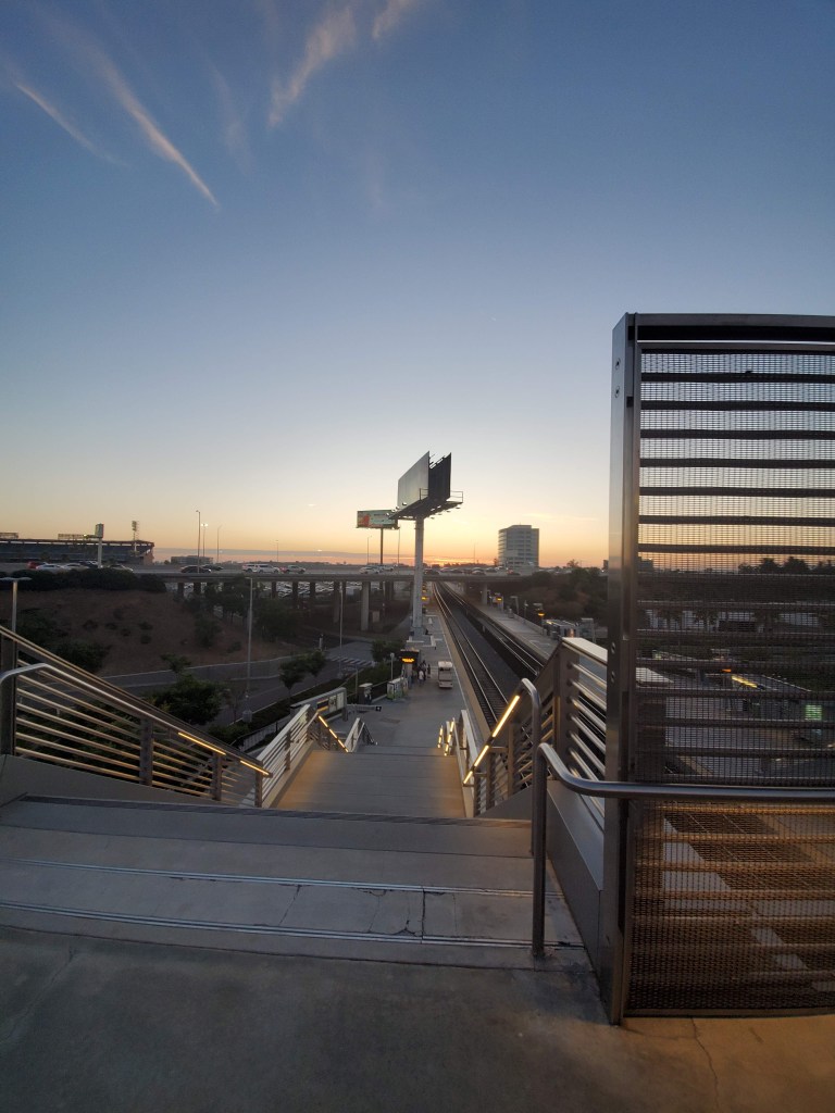 A stair case leading down to a train platform. The sun is setting on a highway. 